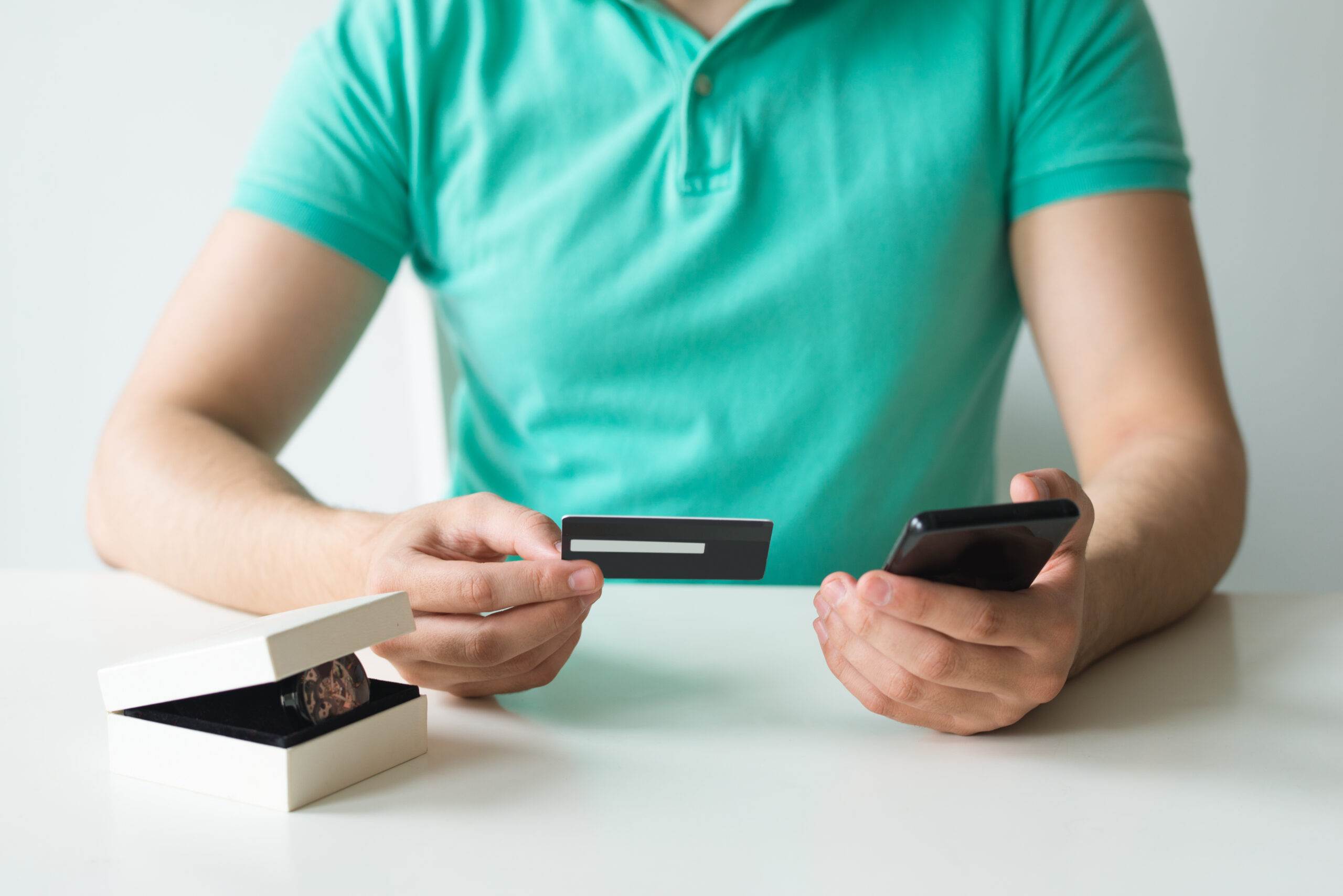 Closeup of buyer holding credit card and smartphone. Person making online payment at table with new watch in box. Online shopping and purchase concept. Cropped front view.