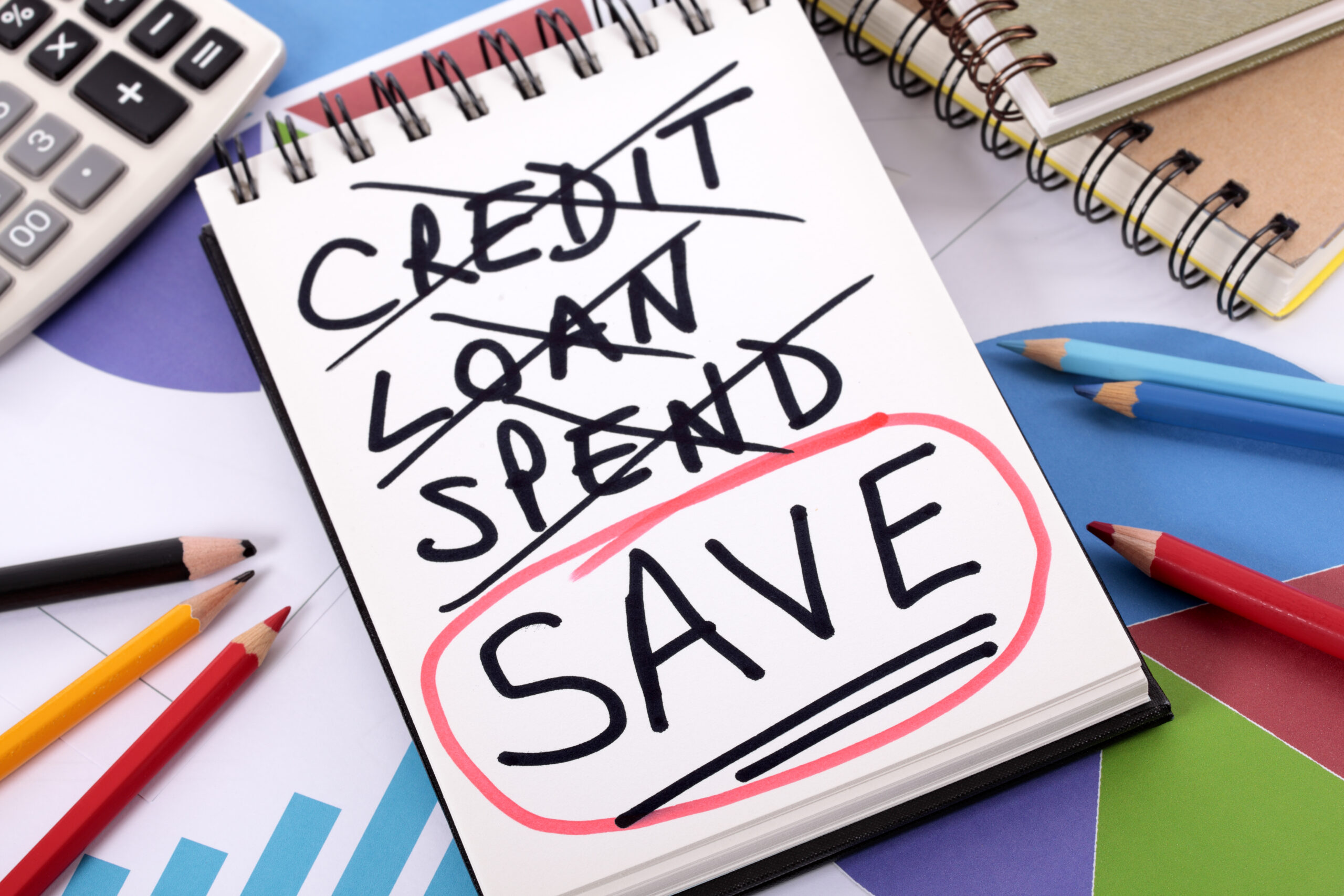 The word Save circled in red below a list of spending written on a notepad surrounded by pencils, graphs, books and calculator.